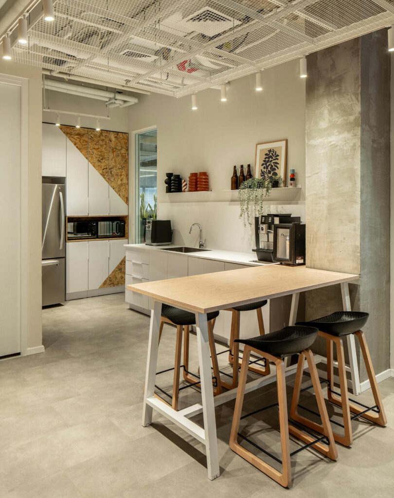 Cuisine de bureau moderne avec une table en bois, quatre tabourets et armoires. Cette chambre dispose d'une cafetière, d'un four micro-ondes, d'un réfrigérateur et de plantes décoratives. Le plafond présente des tuyaux et des lumières apparents.