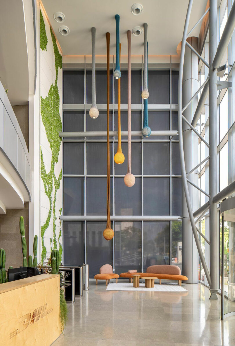 Lobby moderne avec de hautes fenêtres, des sculptures suspendues et un coin salon orange. Des plantes de cactus bordent le côté.