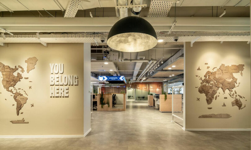 Modern office entrance with beige walls, world map art, and "You Belong Here" text. Large black ceiling light above open workspace with cubicles and glass meeting rooms.