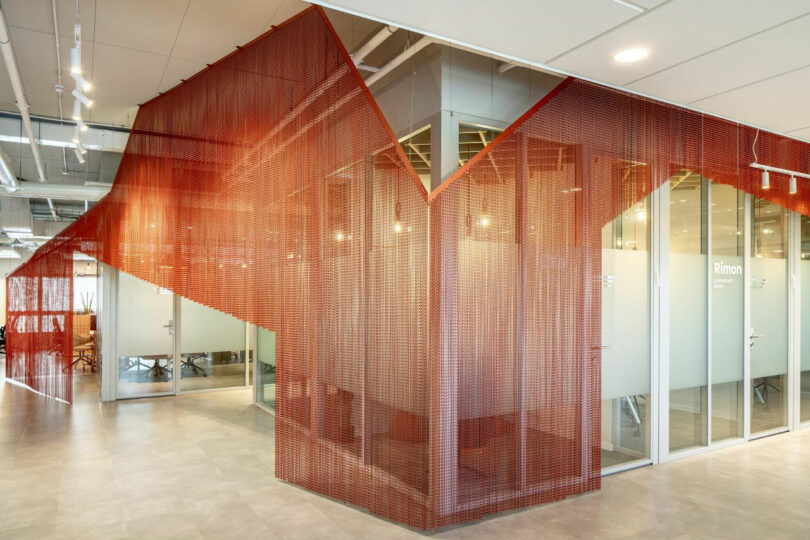 Couloir de bureau moderne avec cloison en maille rouge et salles de réunion aux parois de verre. Les plafonniers éclairent l’espace.