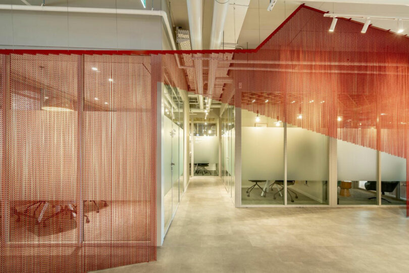 Modern office space with frosted glass walls, open ceilings, and red mesh partition. Minimalist design with neutral flooring.