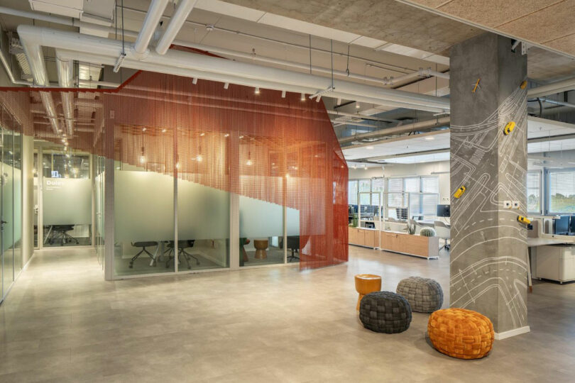 Modern office space with glass partitions, orange mesh panel, and large window. Three woven poufs sit near a pillar with abstract designs and model cars. Ceiling pipes are exposed.