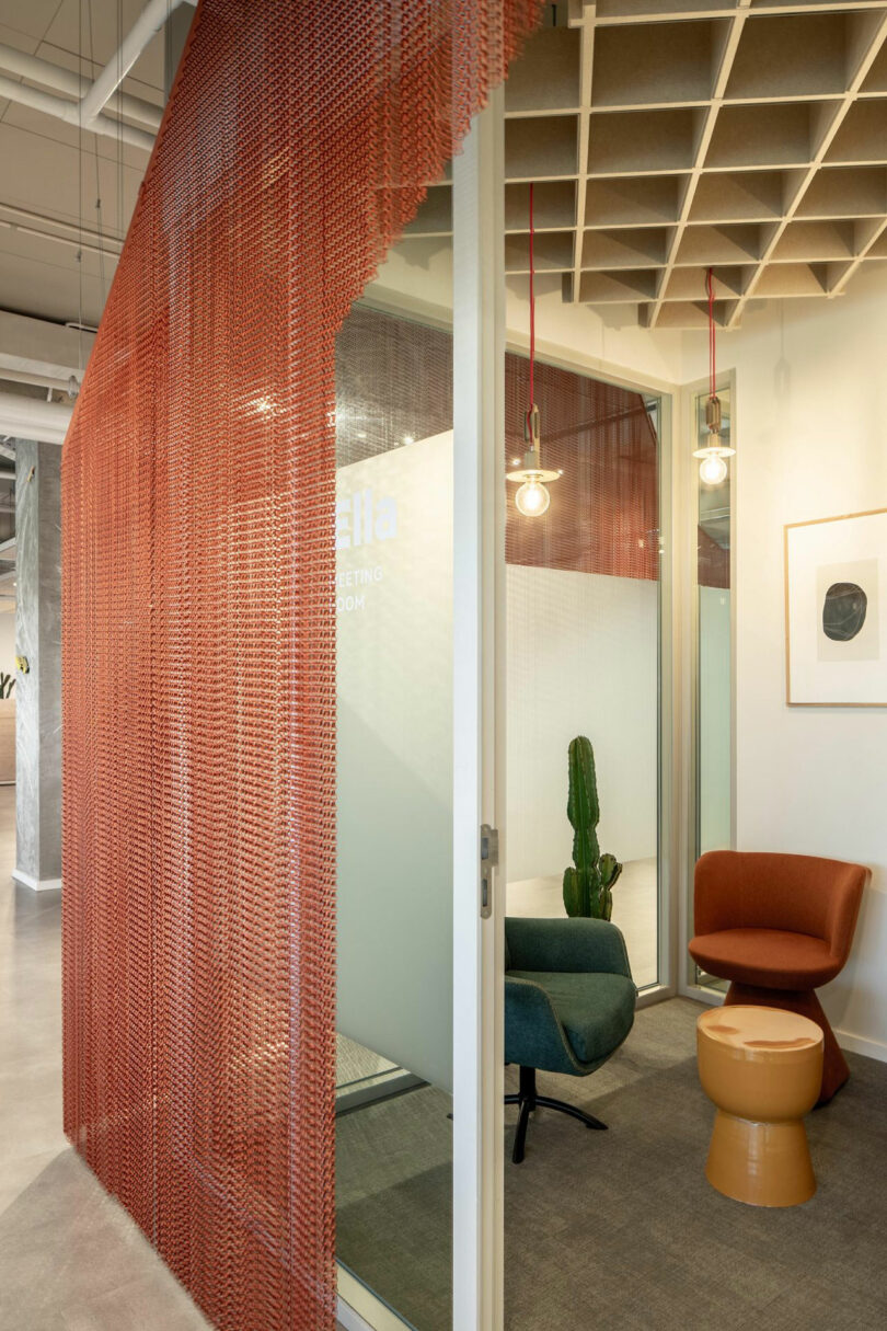 Modern office nook with a red mesh curtain, glass walls, a green chair, an orange chair, a small round table, a cactus, and pendant lights.