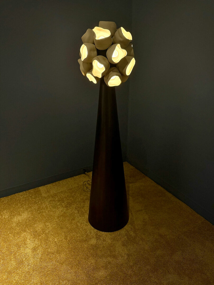 A modern floor lamp with an abstract design, featuring glowing geometric shapes atop a tall, dark cone-shaped stand on a carpeted floor.