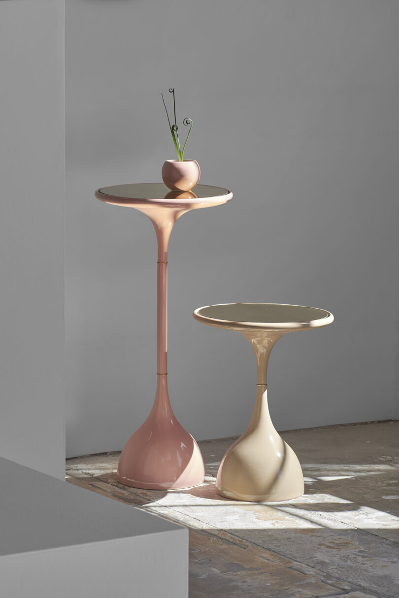 Two sleek, modern pedestal tables with glossy finishes; one tall and pink, the other shorter and beige. A small vase with green stems sits on the taller table, reminiscent of an Entler Design planter.