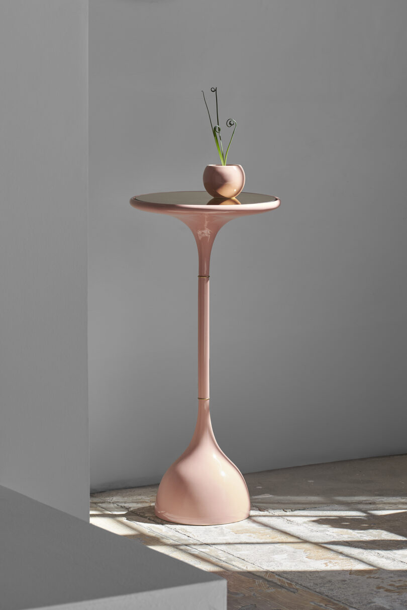 A small pink pedestal table with a glossy surface showcases an Entler Design planter holding a minimalist plant in a round pot, set against a gray wall.