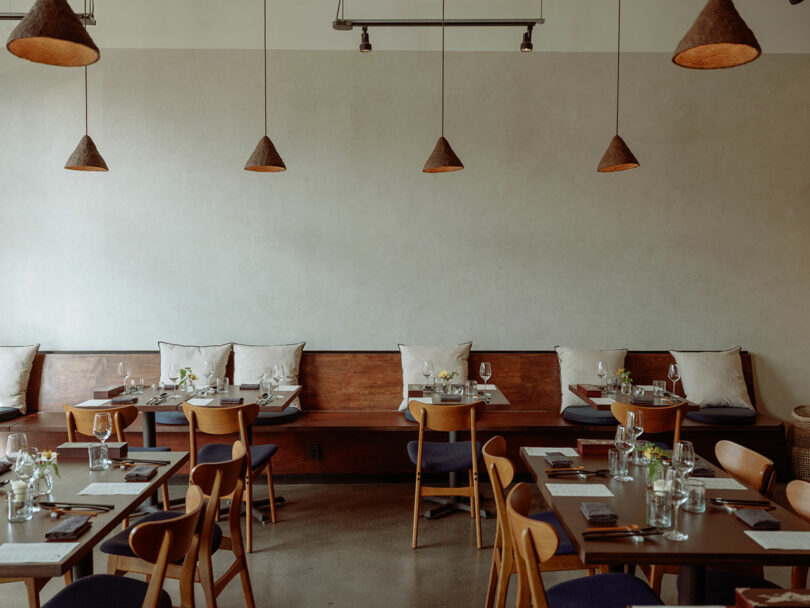 A minimalist restaurant interior designed by Amanda Gunawan features wooden tables and chairs, soft lighting, and simple decor. Each table is elegantly set with glassware and cutlery, reflecting her signature style.