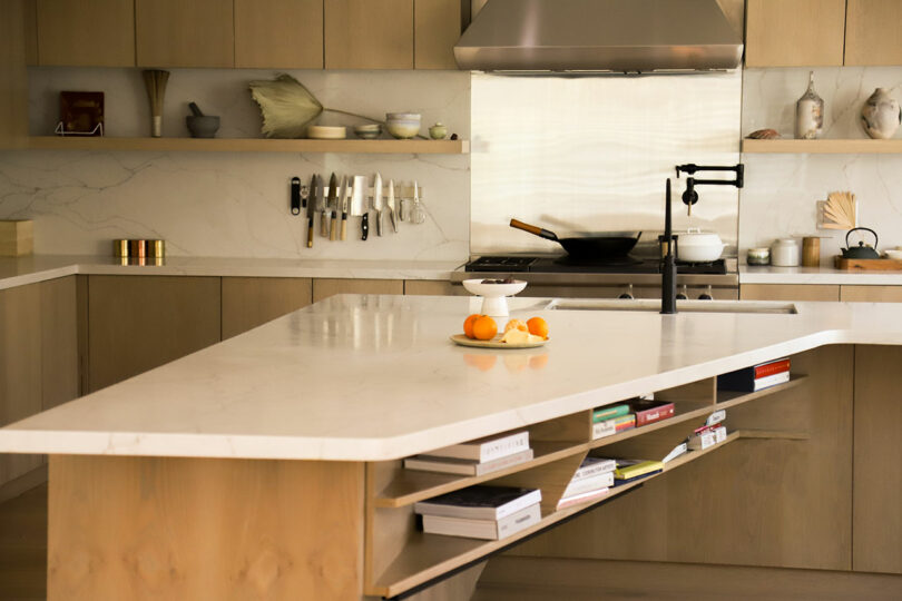 The modern kitchen, with its marble island adorned with fresh oranges and a bowl, reflects Amanda Gunawan's design ethos. Knives neatly line the back wall, complementing the stovetop hood and elegant wooden cabinetry.