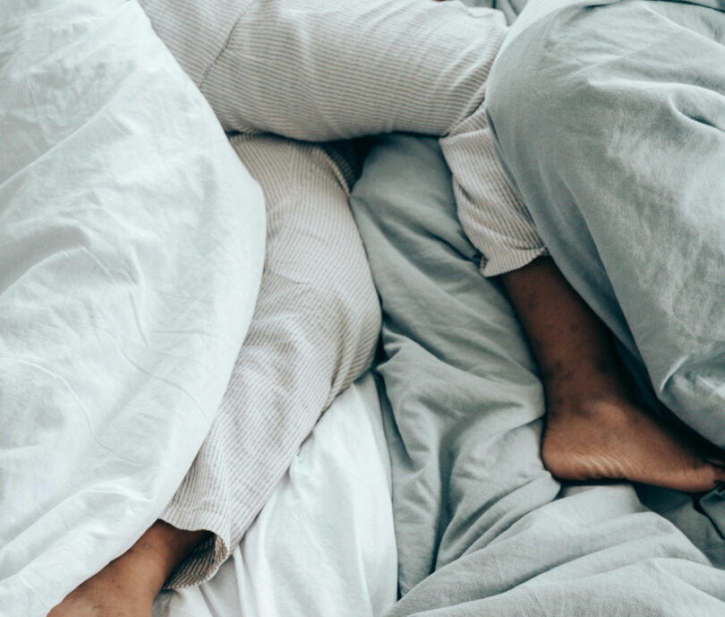 Les pieds et les jambes de deux personnes reposent entrelacés sous des draps blancs et gris, un moment serein qui pourrait inspirer même l'œil artistique de Dan Harden.