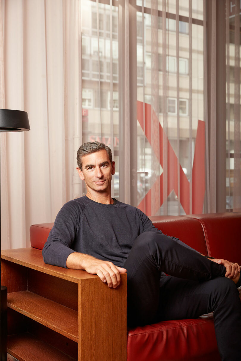 Person sitting on a red couch in a modern room with large windows and wooden furniture.