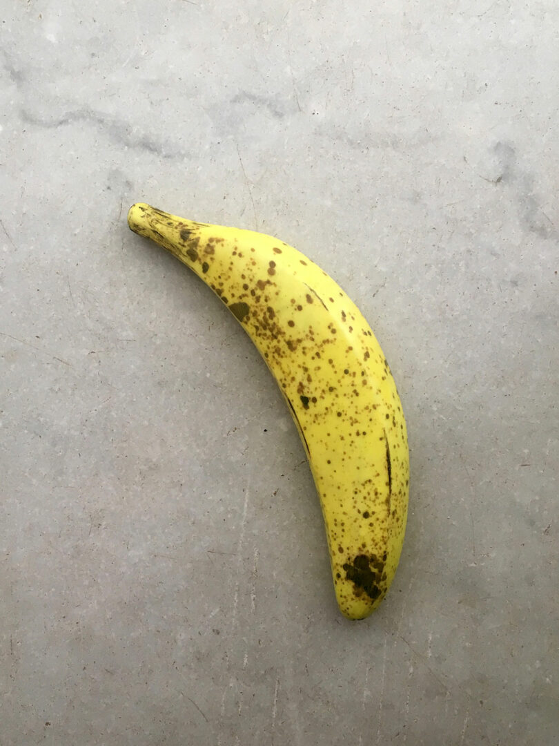 Une banane jaune mûre et mouchetée repose sur une surface gris clair, évoquant l'esthétique minimaliste souvent vue dans les créations de Theodora Alfredsdottir.