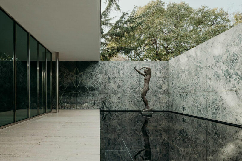 A minimalist outdoor space designed with a Theodora Alfredsdottir-inspired aesthetic features a marble wall, reflective black pool, and a statue. Glass windows on the left and trees in the background complete the serene scene.