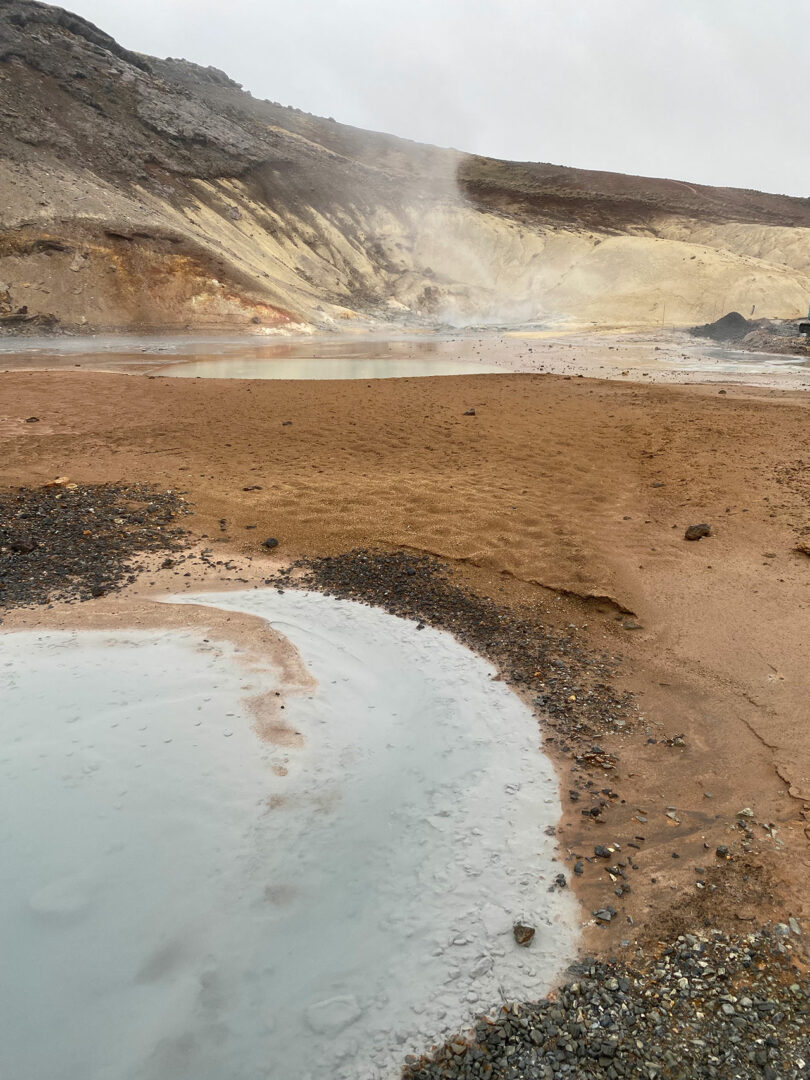 Une zone géothermique naturelle avec des sources chaudes fumantes, des piscines boueuses et un terrain rocheux sous un ciel nuageux reflète le talent artistique synonyme des créations évocatrices de Theodora Alfredsdottir.