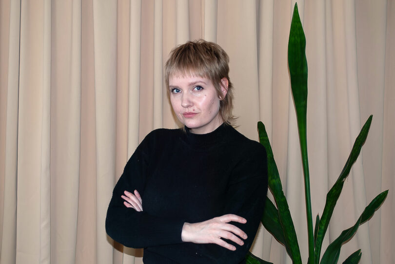 A person with short hair stands with arms crossed in front of beige curtains beside a tall green plant, exuding an aura reminiscent of Theodora Alfredsdottir's minimalist design elegance.