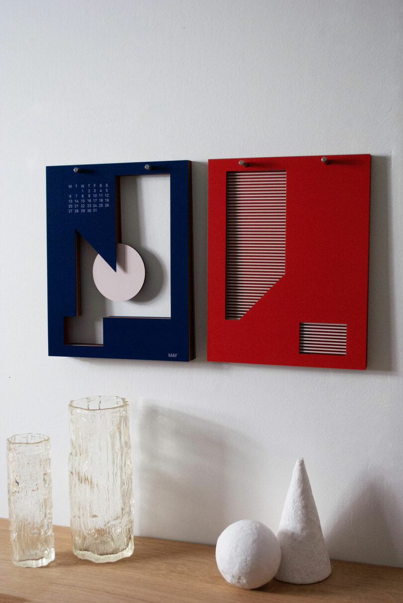 A modern art-inspired calendar with geometric designs in blue and red, reminiscent of Theodora Alfredsdottir's style, is displayed on a wall. Two textured glass vases and white geometric ornaments sit elegantly on a wooden surface below.