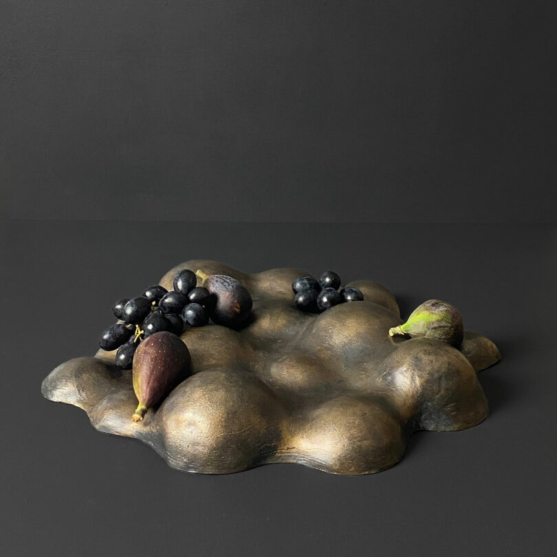 An abstract bronze tray, crafted by Theodora Alfredsdottir, displays figs and grapes beautifully arranged against a dark background.