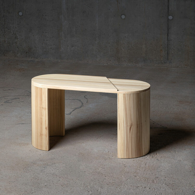 A modern, oval-shaped wooden bench with a cut-out design by Theodora Alfredsdottir rests gracefully against a concrete wall and floor.