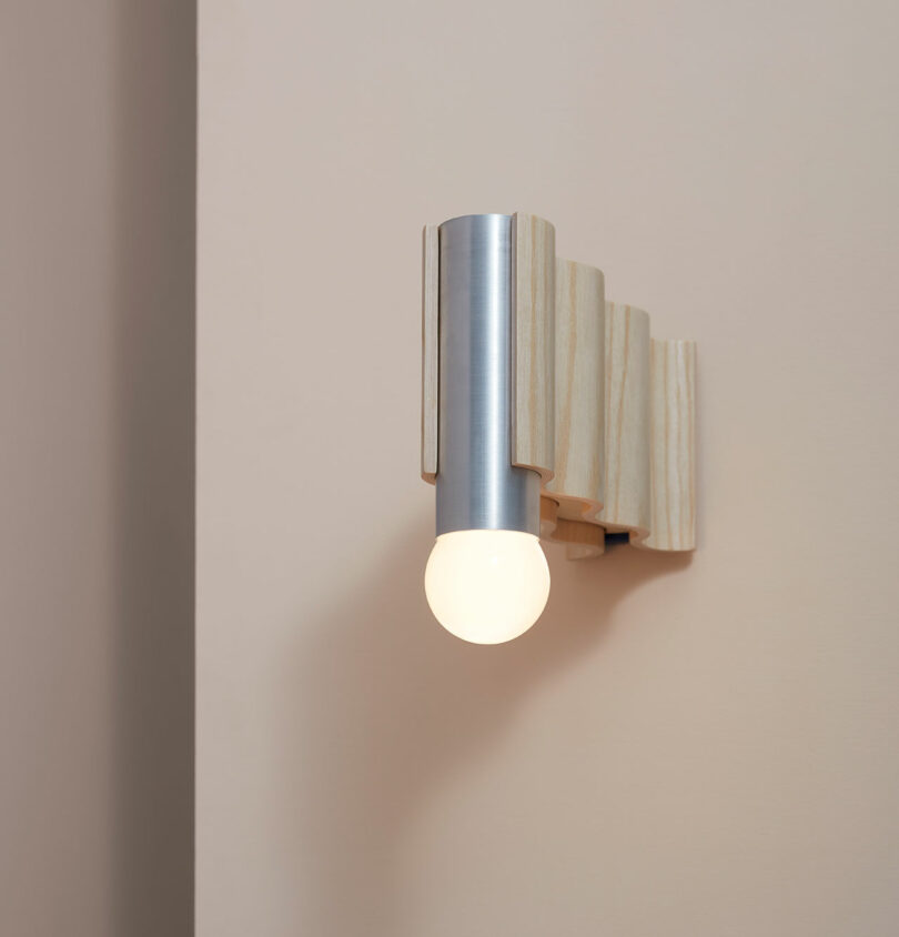 A wall-mounted light fixture, reminiscent of Theodora Alfredsdottir's minimalist designs, features a sleek cylindrical metal base and a glowing bulb set elegantly against a beige background.