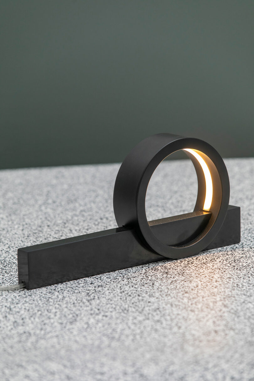 Une lampe circulaire noire moderne, conçue par Theodora Alfredsdottir, présente un anneau ouvert sur une base rectangulaire et repose élégamment sur une surface mouchetée.