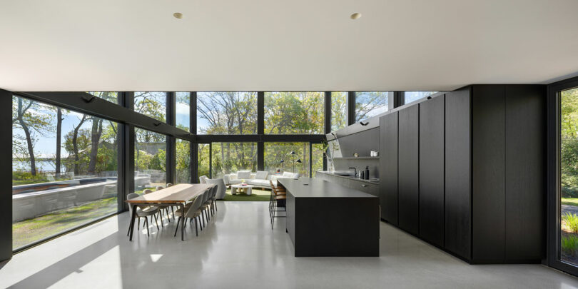 Modern kitchen with floor-to-ceiling windows, a large dining table, black cabinetry, and an island. Trees and a body of water are visible outside.