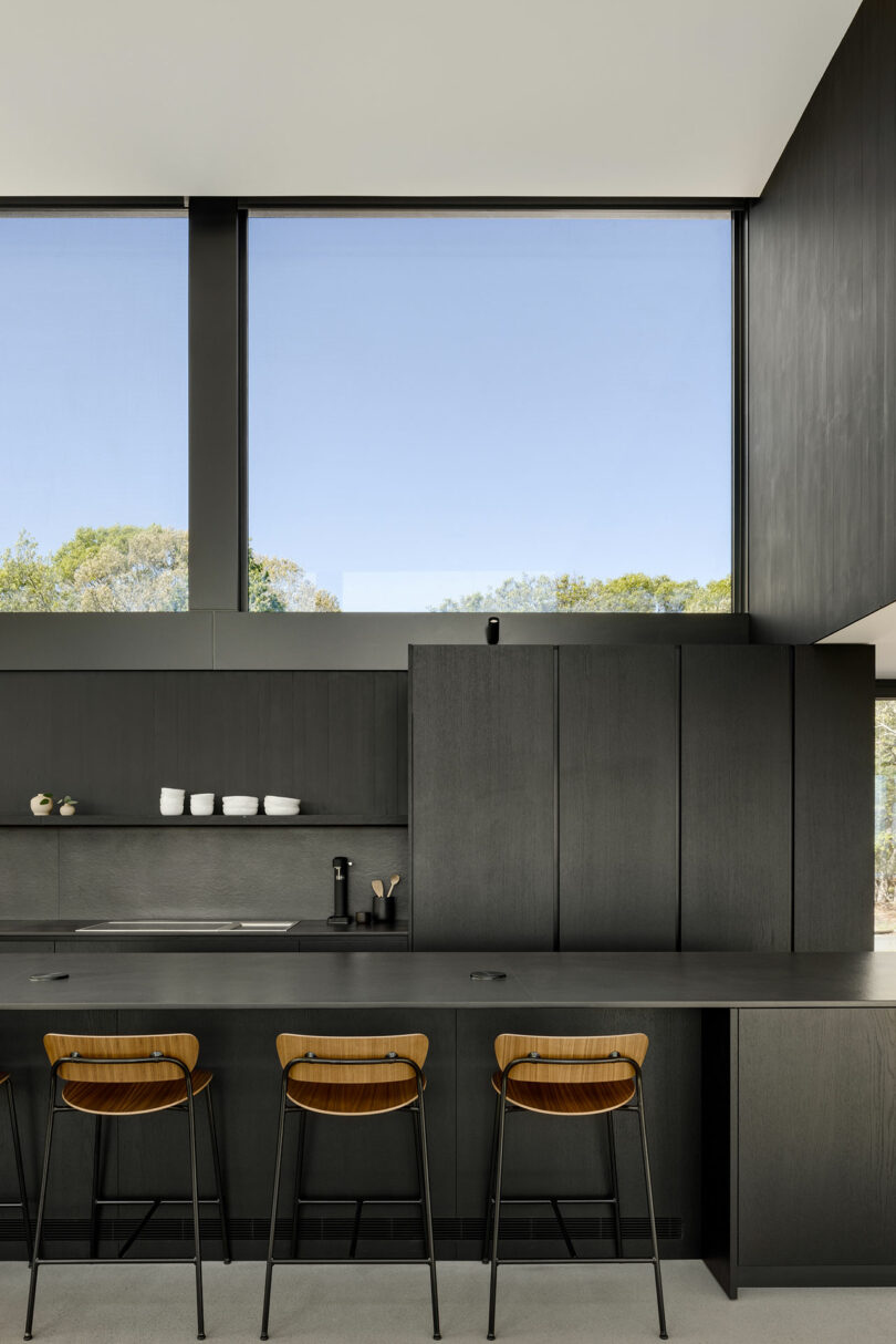 Modern kitchen with dark cabinetry, a long countertop, three wooden barstools, and large windows displaying a clear sky and trees outside.