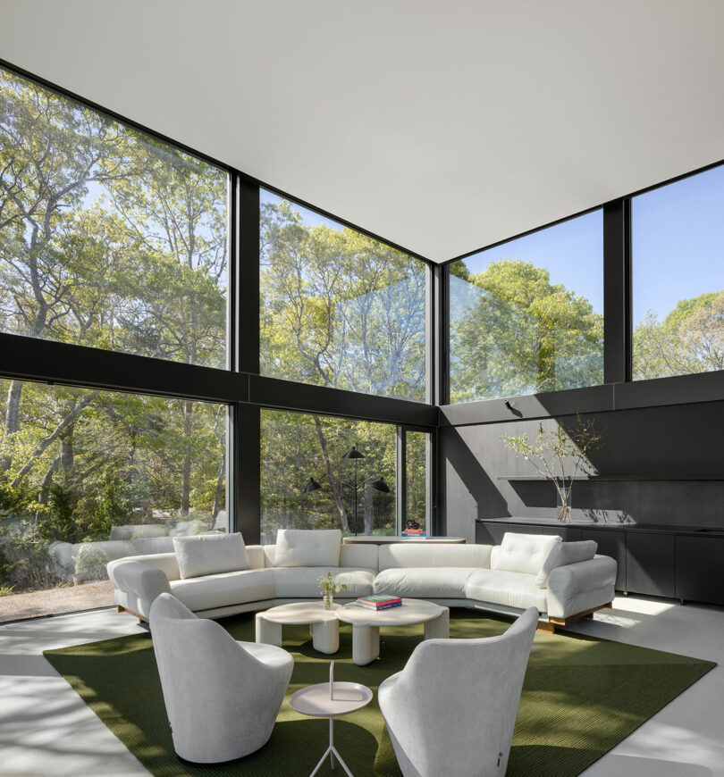 A modern living room with floor-to-ceiling windows, white sofas, green carpet, and minimalist decor, overlooking a lush forest view.