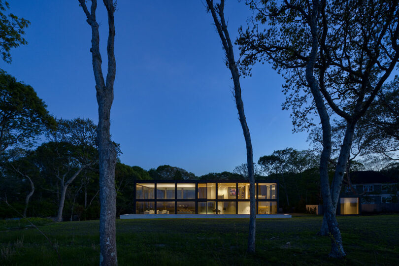 A modern two-story house with large windows is illuminated at dusk, surrounded by trees and a grassy area.