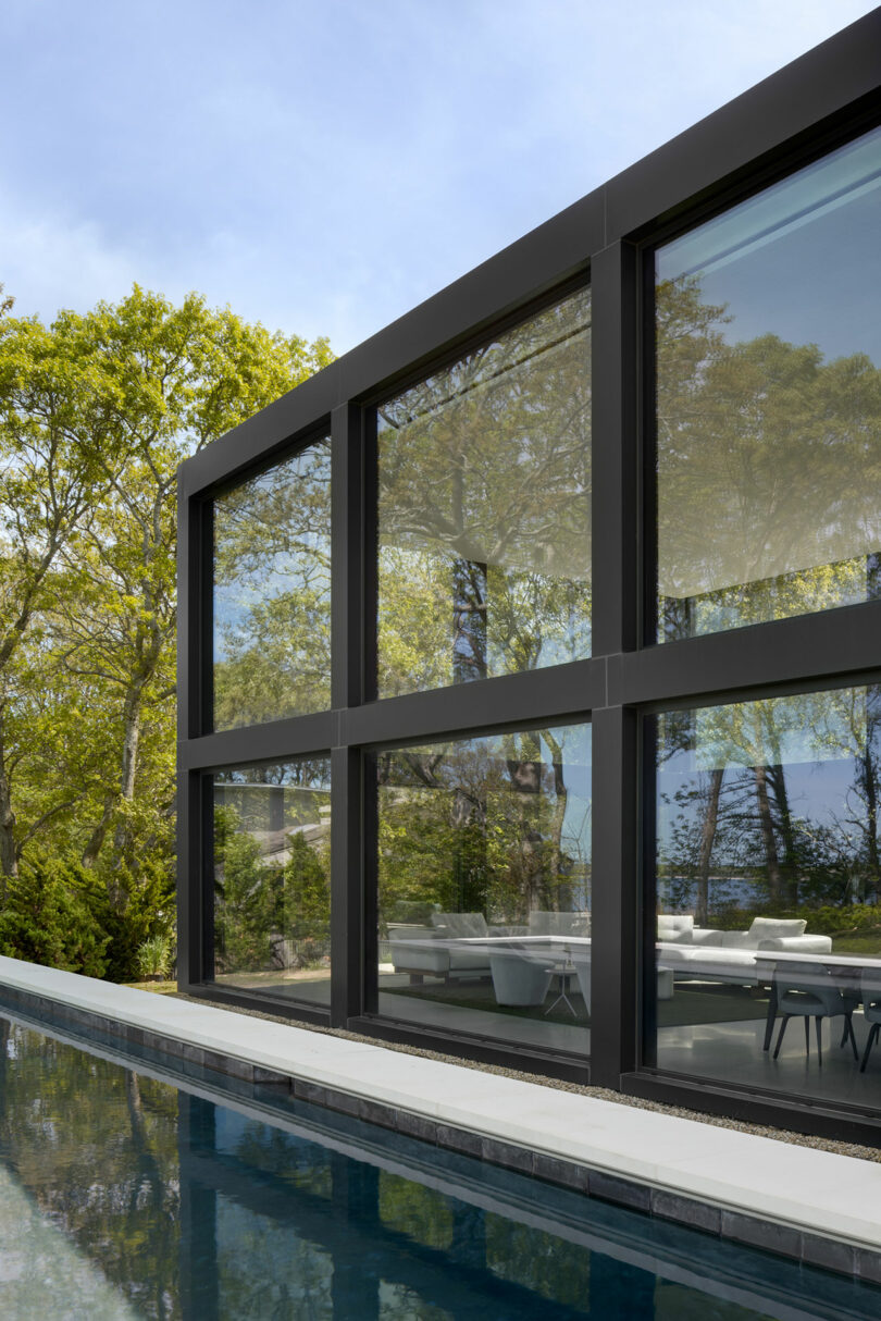 Modern building with large glass windows reflecting trees, adjacent to a swimming pool. The façade is sleek and minimalist, surrounded by greenery.