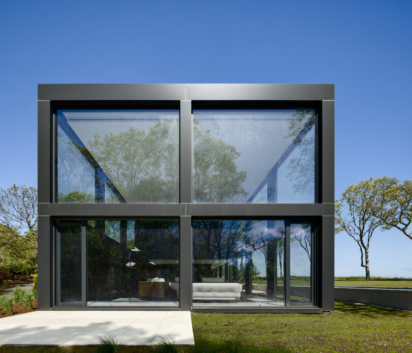 Modern glass and steel house with large windows, surrounded by trees and a clear blue sky.