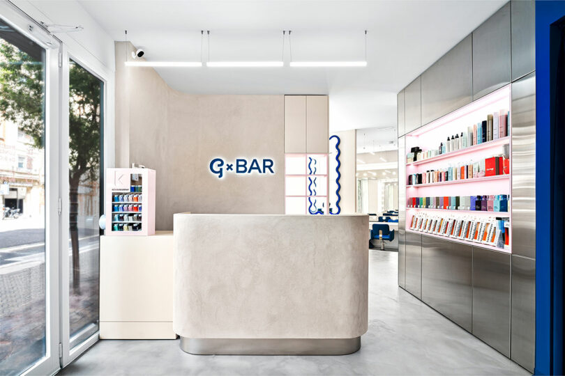 Bright, modern salon interior with a reception desk, illuminated product shelves, and a large window letting in natural light.