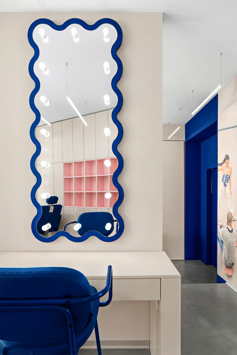 A modern interior with a wavy-edged mirror bordered by lights, a blue chair at a white desk, and a hallway with colorful artwork and open shelving.