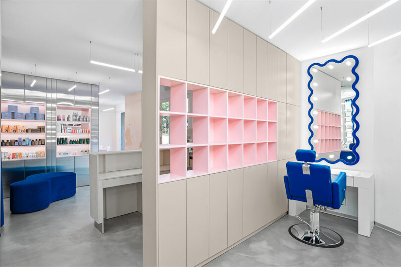 A modern salon interior with a blue styling chair, pink shelves, and a large wavy mirror. The space is well-lit with a minimalist design and displayed hair products in the background.