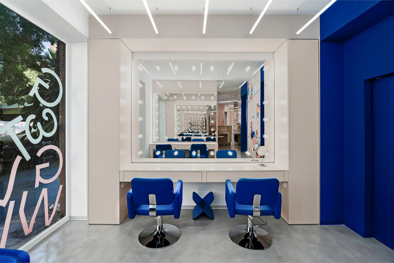 A modern hair salon interior with blue chairs, a large mirror, and bright overhead lighting.