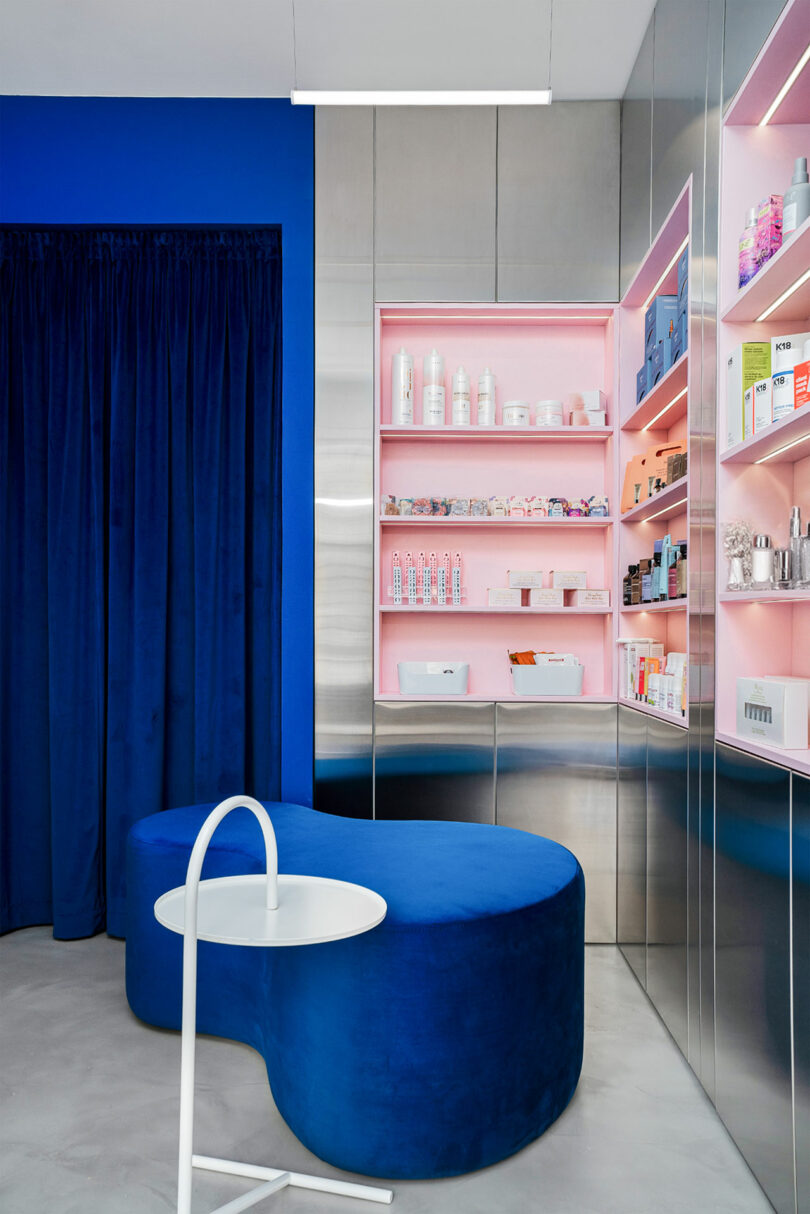 Modern interior with a blue velvet bench, stainless steel cabinets, and shelves displaying various products. A white round table is in front of the bench, and a dark blue curtain is in the background.