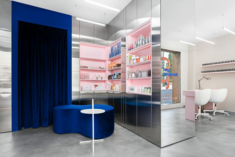 Modern beauty salon interior with metallic walls, shelves displaying various products, a blue velvet seating area, and a manicure station with white chairs.