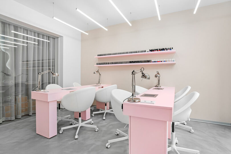 A modern nail salon with pink manicure tables, white chairs, and shelves displaying nail polishes.