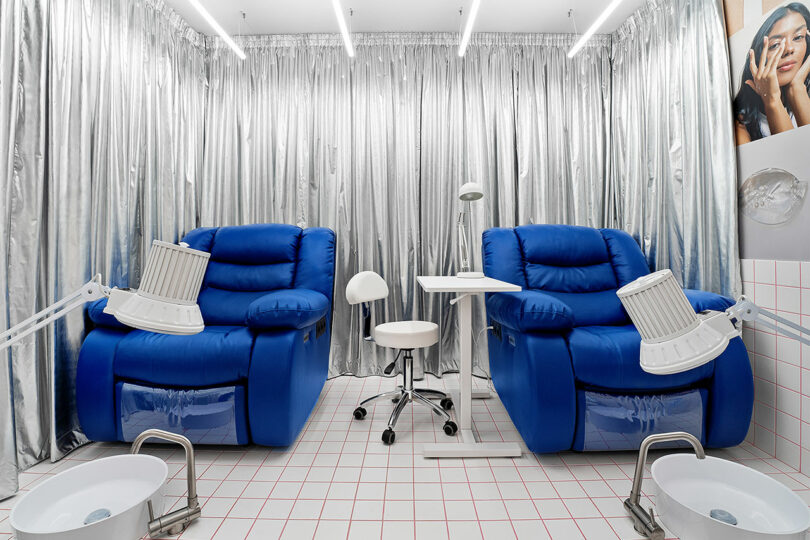 A futuristic salon setup with two blue reclining chairs, drying lamps, foot baths, a small table, and a stool, all surrounded by metallic silver curtains.