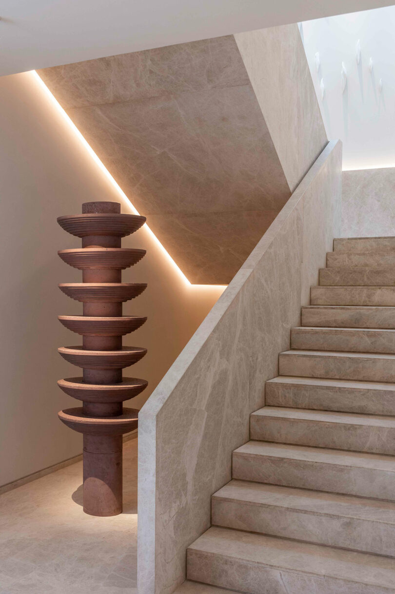 A modern, minimalist staircase with beige marble steps and walls. On the left, a stacked circular wooden sculpture stands vertically. Soft lighting highlights the design.