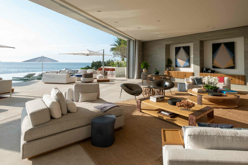 Spacious modern living area with beige sofas and wooden tables, opening to an outdoor patio with poolside chairs, umbrellas, and ocean views. Large art pieces adorn the walls.