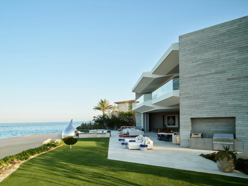 Modern beachfront house with expansive patio, outdoor seating, and grill. Large lawn and metal sculpture in the foreground. Ocean and clear sky in the background.