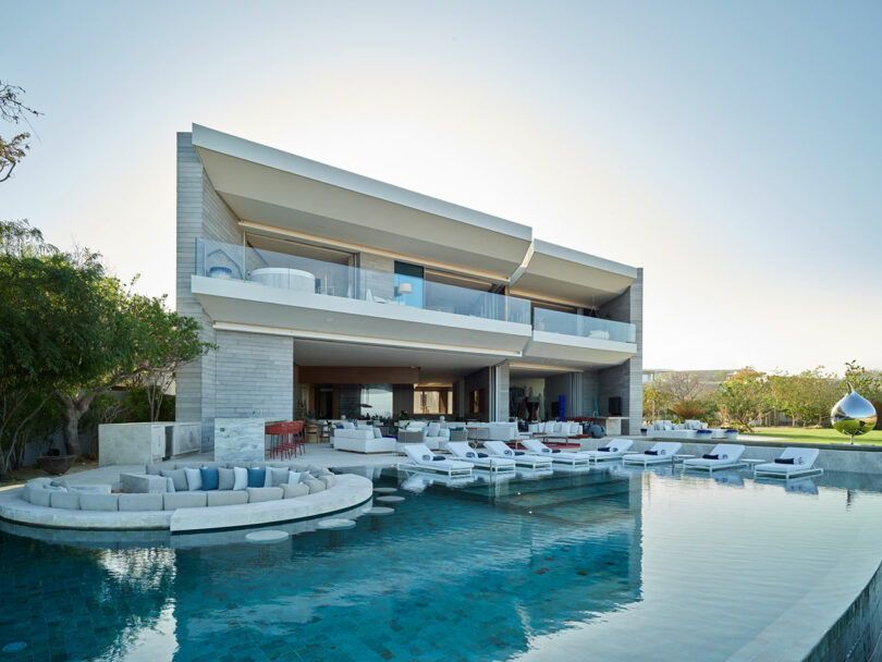 Modern two-story house with large glass windows, open balconies, and an expansive pool surrounded by sun loungers and a round seating area. Trees and a unique outdoor chair are visible.