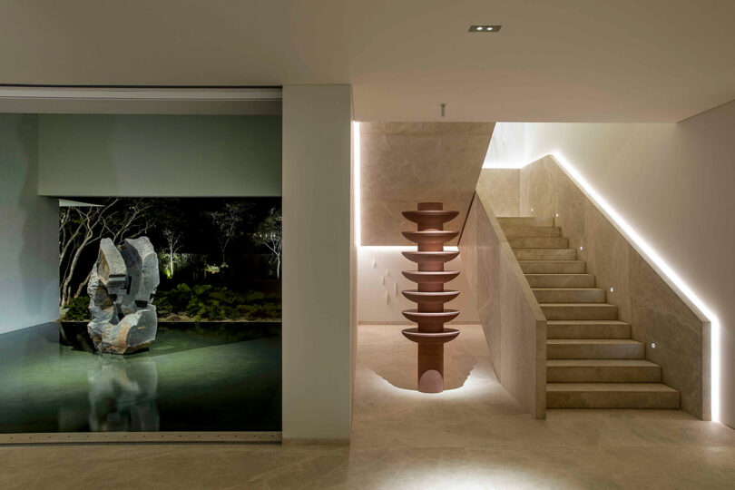 Minimalist interior featuring a lit staircase, water feature with sculpture, and textured stone finishes.