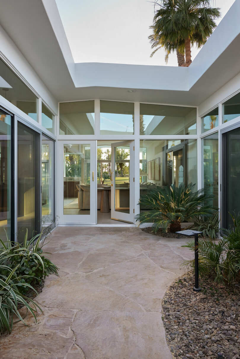 Modern courtyard with glass doors and windows, surrounded by plants and trees, leading to an indoor space.