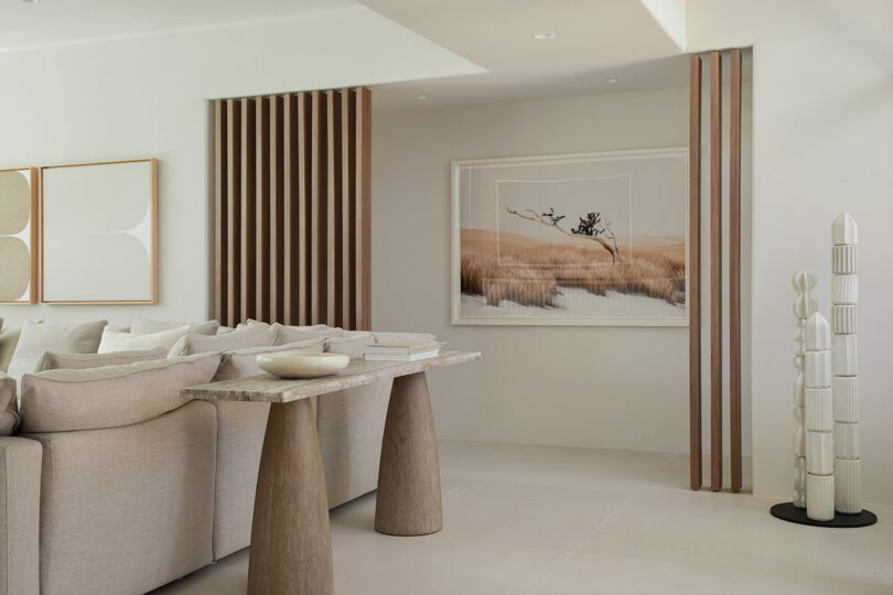 Minimalist living room with beige couch, abstract wall art, and decorative vertical wood slats. A table with a ceramic bowl and books is in the foreground.