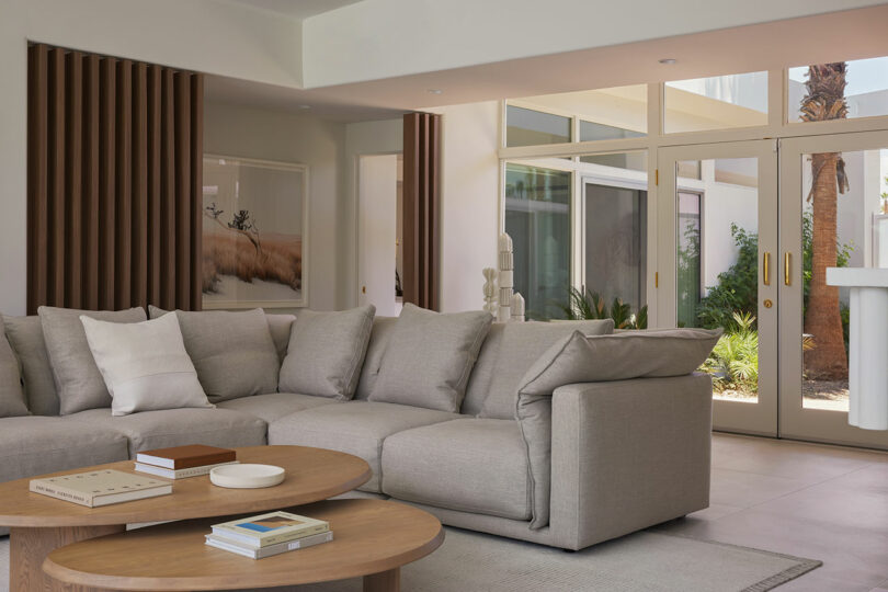 Modern living room with a large gray sectional sofa, round wooden coffee tables with books, and floor-to-ceiling windows showcasing a view of a garden.