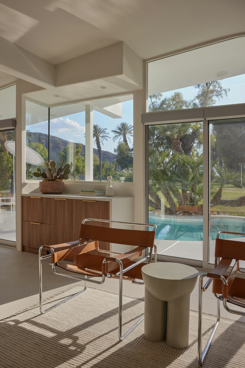 Modern living room with large windows, brown leather chairs, and a view of a pool and palm trees outside. A beige rug covers the floor, and a small table is between the chairs.