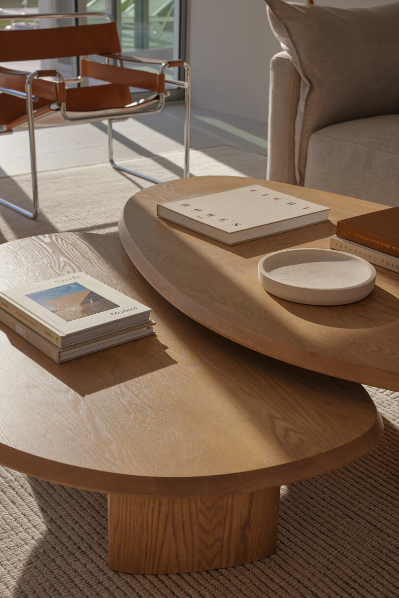 Wooden coffee table with stacked oval tops, featuring books, a round dish, and a tan chair in the background. A sofa is partially visible on the right.