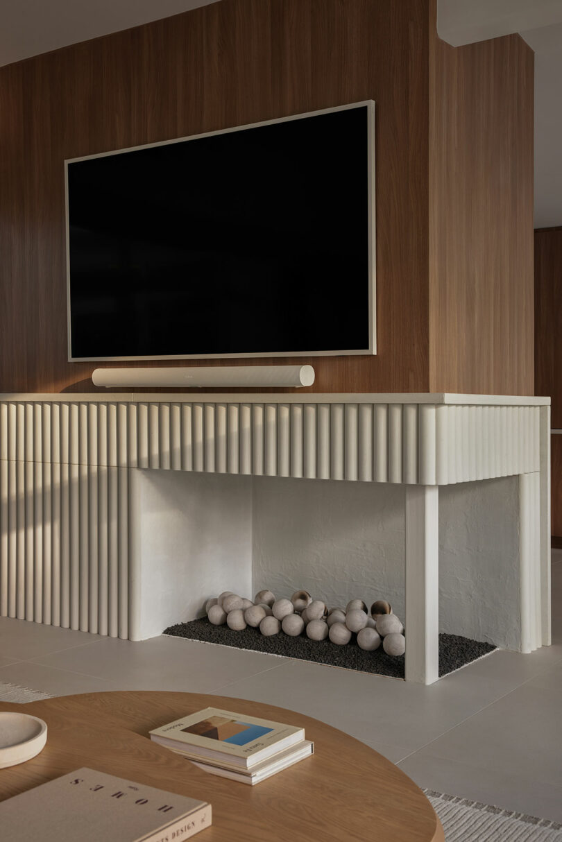 Modern living room with a flat-screen TV mounted above a minimalist fireplace. The fireplace features a textured white facade and decorative spheres inside. A wooden coffee table is in the foreground.