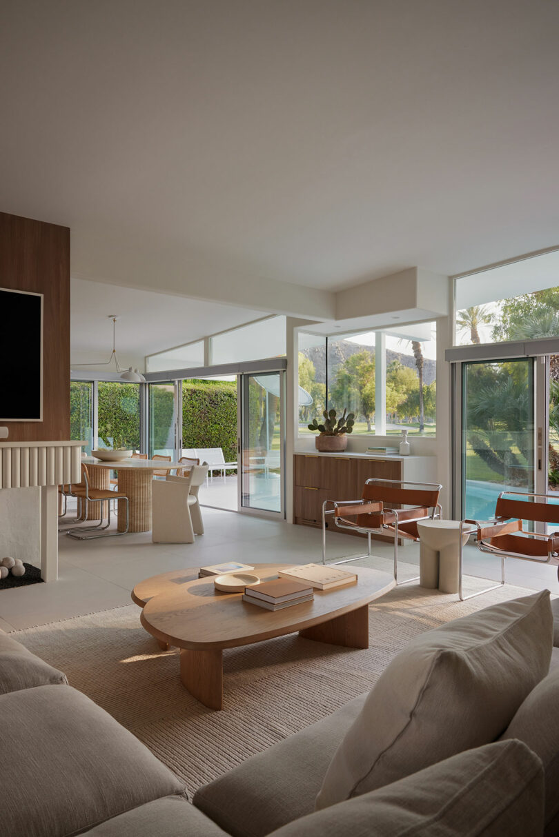 Modern living room with neutral tones, featuring a beige sofa, wooden coffee table, and large windows overlooking a garden and pool.