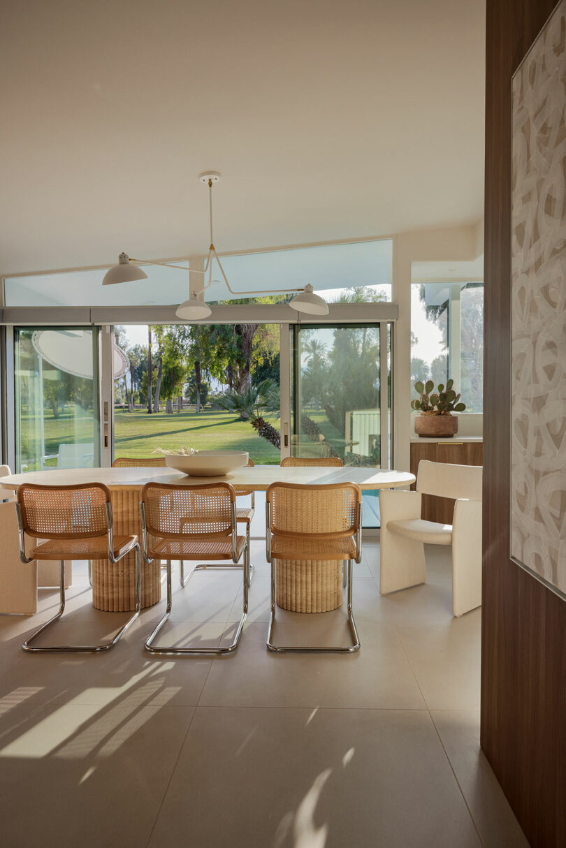 Modern dining area with wicker chairs, a rectangular table, and large sliding glass doors opening to a garden view. Natural light fills the space, highlighting minimalist decor and a potted plant.